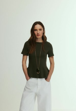 Woman stands with hands in pockets wearing dark green short-sleeve top, long pendant necklace, and white wide-leg pants against light background.