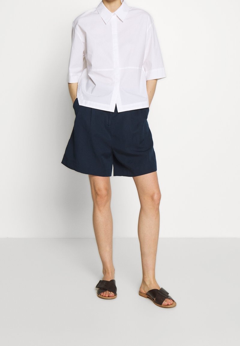 Chemise blanche courte à boutons avec manches courtes, associée à un short bleu marine. Sandales marron à enfiler avec un design à bride croisée.