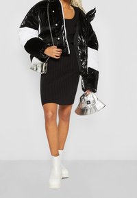 Femme portant une robe noire côtelée et une doudoune brillante noire et blanche, tenant un chapeau argenté et un petit sac à main métallique, chaussée de bottes blanches.