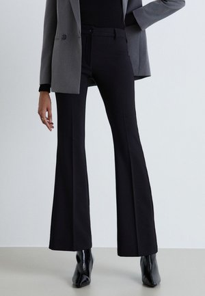 Personne portant un pantalon noir évasé, des bottes noires, un haut noir et un blazer gris, debout avec une main sur la hanche et l'autre détendue.