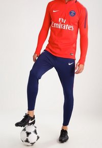 Maillot de football rouge à manches longues avec fermeture éclair à demi-hauteur et logo blanc "Fly Emirates", associé à un legging bleu marine et des chaussures de football noires, debout sur un ballon de football.