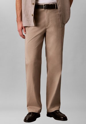 Man die beige broek, een licht beige open overhemd, zwarte riem en zwarte nette schoenen draagt, staande tegen een neutrale achtergrond.
