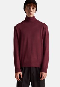 Maglione a collo alto bordeaux realizzato in materiale morbido e sottile, con una vestibilità aderente e polsini a coste. Indossato con pantaloni neri larghi.