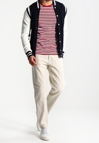 Hombre caminando con una chaqueta varsity azul marino y blanca, camiseta a rayas rojas y blancas, pantalones beiges y zapatillas blancas sobre un fondo blanco.