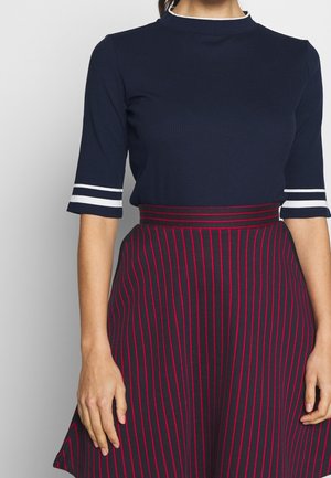 Femme portant un haut bleu marine avec des rayures blanches sur les manches et une jupe bleu marine avec des rayures rouges verticales et une ceinture rayée.