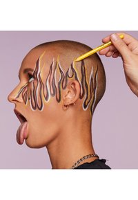 Une tête chauve présente un maquillage du visage audacieux en forme de flammes, aux couleurs marron, jaune et blanche. Une main applique un crayon jaune sur le dessin devant un fond lilas.