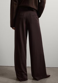 Pantalones de pierna ancha en un tejido burdeos profundo, con dos bolsillos traseros y una textura suave, combinados con un suéter marrón.