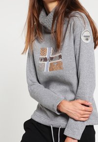 Grijze trui met een colnek en lange mouwen. Heeft bruine, beige en witte chevronborduursels aan de voorkant en een embleem op de mouw.