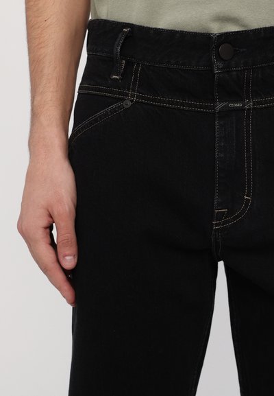 Vaqueros negros con un diseño ajustado, que presentan costuras en contraste, un cierre con botón y una pequeña etiqueta de la marca cerca de la cintura.