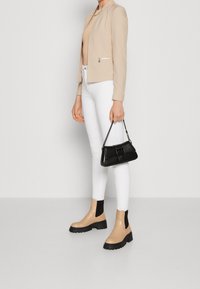 Beige tailored jacket with a zipper accent, paired with white skinny pants and beige ankle boots. Holding a black handbag with a chain strap.