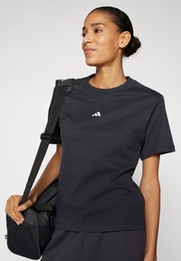 T-shirt de sport noir à manches courtes en tissu lisse, avec un petit logo blanc sur la poitrine et une coupe décontractée. Sac à bandoulière noir tenu à la main.