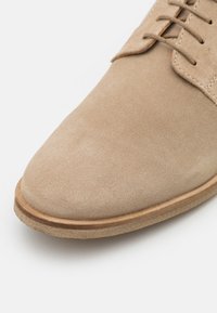Chaussure en daim beige avec une texture lisse, présentant un bout rond et un design classique à lacets. La semelle est plate et fabriquée en caoutchouc de couleur claire.