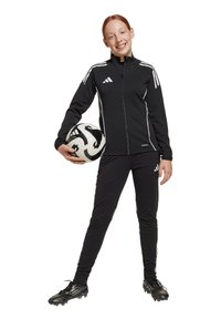 Zwarte adidas trainingspak met witte strepen, slim fit ontwerp, rits aan de voorkant, gecombineerd met een zwart-wit voetbal en zwarte voetbalschoenen.