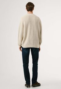 Man standing facing away wearing a cream knit sweater, dark blue jeans, and brown sneakers against a plain light background.