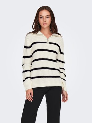 Femme portant un pull en maille blanc avec des rayures horizontales noires et un col à zip partiel, assorti à un pantalon noir, debout sur un fond blanc.