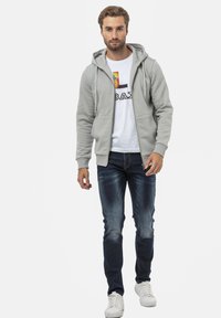 Veste à zip grise avec poches avant doubles portée sur un t-shirt blanc avec un graphisme coloré, associée à un jean bleu délavé et des baskets blanches.