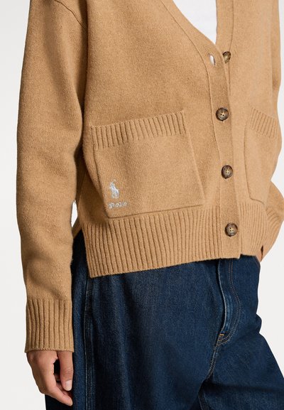 Camel-colored knitted cardigan with a V-neck, featuring a button closure, two front pockets, ribbed cuffs, and an embroidered logo on the pocket.