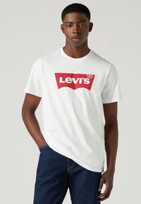 Hvid bomulds t-shirt med et fremtrædende rødt Levi's-logo i et buet rektangulært design, parret med mørkeblå denim jeans.