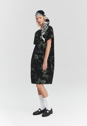 Mujer con vestido negro de flores, calcetines blancos, zapatos negros y pañuelo en la cabeza con patrón en blanco y negro, de pie contra un fondo liso.