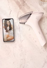 Philips Lumea ontharingsapparaat in het wit met zilveren accenten, naast een telefoon die een gebruikersinterface toont op een marmeren oppervlakte.