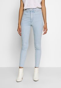 Žena v světle modrých úzkých džínách s vysokým pasem, bílém bezrukávovém topu a bílých kotníkových botách stojí proti jednolitému pozadí.