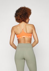 Narancssárga sportmelltartó keresztpántokkal és fehér Nike logóval; magas derékú olíva leggings-szel párosítva, sima textúrával és illeszkedő kialakítással.