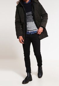 Ciemnozielona parka z futrzanym kapturem, nałożona na niebiesko-szary sweter w paski, w połączeniu z czarnymi dopasowanymi spodniami i czarnymi botkami.