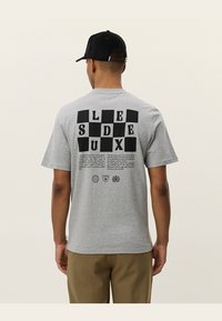 Světle šedé bavlněné tričko s černým grafickým potiskem písmen na zádech, které obsahuje design šachovnice a další text pod ním.