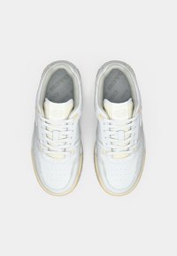 Baltos odinės sportbačiai su lygiu paviršiumi, kurių priekyje yra perforuota dalis, baltos raišteliai ir šviesios spalvos gumine dūpliu. Matomos pilkos vidpadžio dalys.