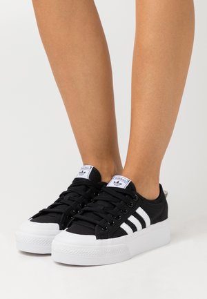 Piernas que llevan zapatillas de Adidas en blanco y negro de tipo bajo con suelas blancas gruesas, de pie sobre un fondo claro.