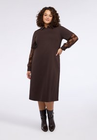 Brązowa sukienka z krótkimi rękawami, ozdobiona przezroczystymi koronkowymi akcentami na mankietach, klasycznym kołnierzykiem i prostym krojem. Stylizowana z kozakami za kolano.