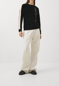 Persona che indossa un top a maniche lunghe nero con strisce crema e borgogna sulle maniche e pantaloni bianchi a gamba larga con scarpe nere.