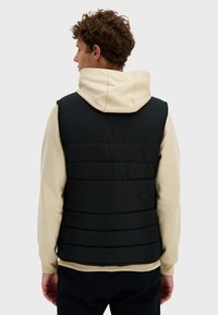 Czarna ocieplana kamizelka z pikowanym wzorem, noszona na jasnoszarej bluzie z kapturem. Posiada wysoki kołnierz oraz elastyczne akcenty w talii i mankietach.