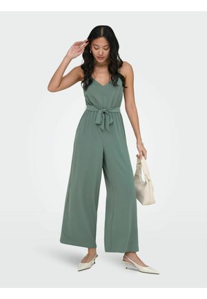 Kobieta w bezrękawnikowym kombinezonie o szerokich nogawkach w kolorze szałwiowej zieleni z wiązaniem z przodu, trzymająca kremową torebkę i nosząca kremowe sandały na obcasie z paskiem z tyłu.