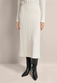 Knit cream pleated skirt with a high waistband, paired with pointed black heeled boots. Smooth texture, mid-length design, and vertical lines.
