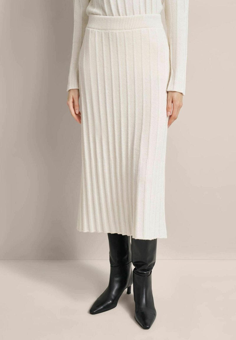 Knit cream pleated skirt with a high waistband, paired with pointed black heeled boots. Smooth texture, mid-length design, and vertical lines.