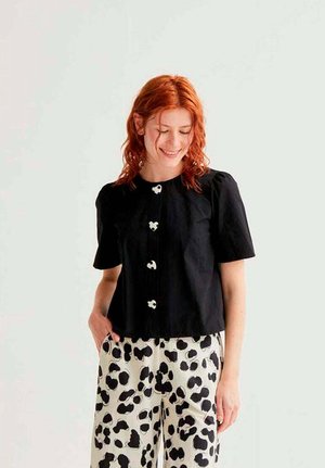 Mujer con cabello rojo lleva una blusa negra de manga corta con detalles de botones y pantalones blancos con un patrón de manchas abstractas negras.