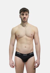 Mutande nere a slip con marchio bianco, caratterizzate da un design a vita bassa. Il modello è in piedi, con uno sfondo neutro e accessori minimi.