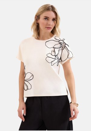 Woman with blonde hair wears a white short-sleeve top with black floral line designs and black pants against a plain background.