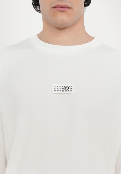 Vit bomull t-shirt med en liten rektangulär svart och vit etikett med siffror och en cirkulär grafisk design på bröstet.