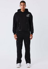 Zwarte hoodie met voorzak, voorzien van witte verfspetters en grafische print. Gecombineerd met zwarte broeken, ook met verfdetails en zakken.