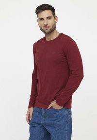 Bordeauxrood shirt met lange mouwen van zachte stof, met een ronde halslijn en een klein logo op de borst. G gedragen met blauwe spijkerbroek.