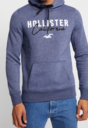 Mężczyzna w niebieskiej bluzie z kapturem Hollister California z przednią kieszenią i ciemnoniebieskich dżinsach na jednolitym tle.