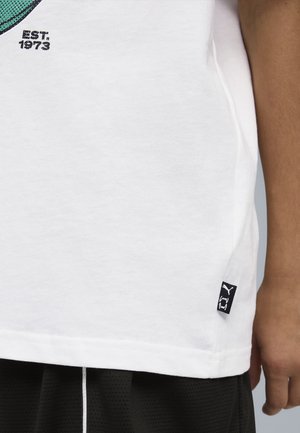 Biała koszulka z nadrukiem "EST. 1973" w pobliżu ramienia i małą czarną metką z logo Puma przy brzegu, noszona z czarnymi sportowymi szortami.