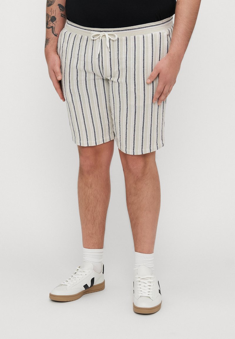 Shorts rayés crème avec des lignes navy, dotés d'une taille à cordon de serrage ajustable. Associés à des baskets blanches et des chaussettes côtelées.