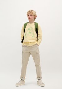 Geel sweatshirt met groene doodskopgraphic, beige cargo broek, groene rugzak en witte sneakers. Casual, comfortabele stijl en pasvorm.