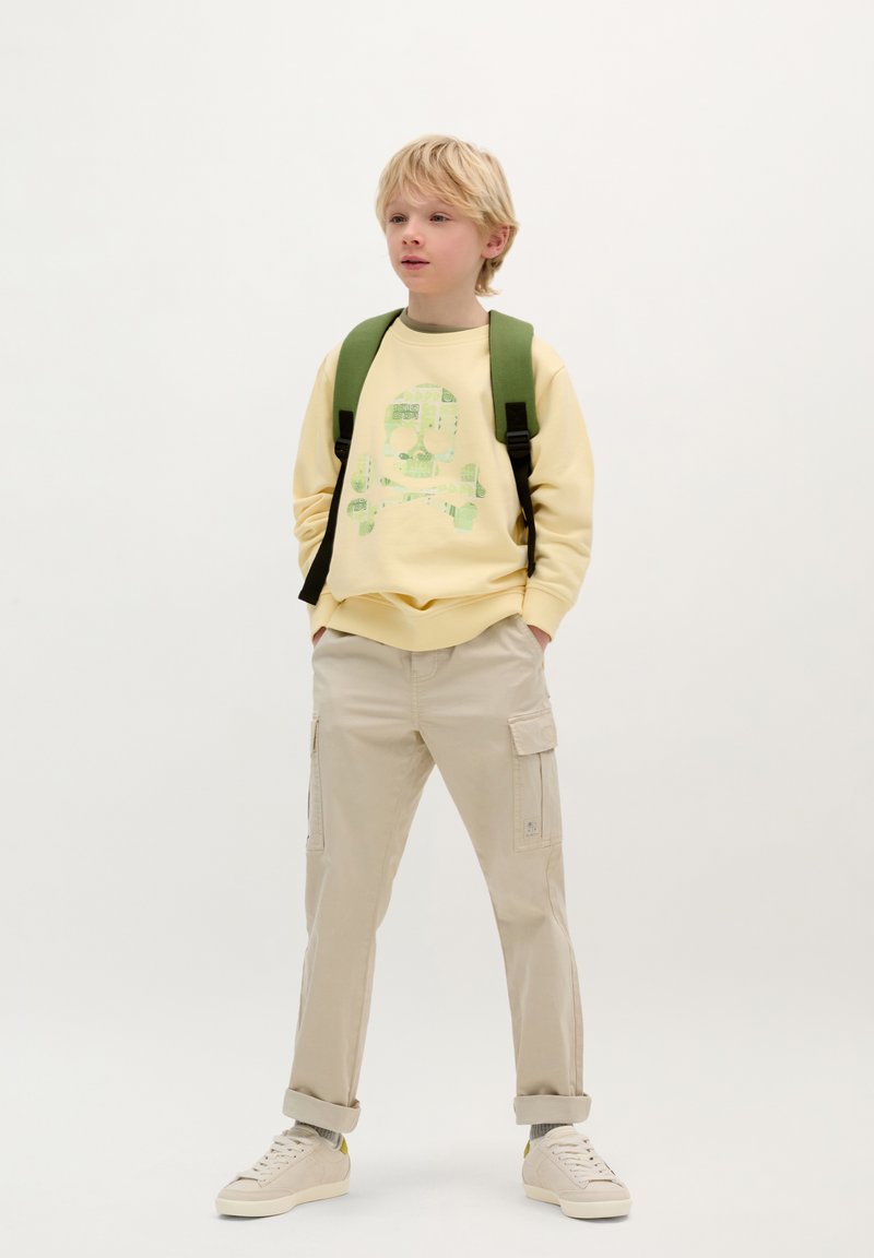 Geel sweatshirt met groene doodskopgraphic, beige cargo broek, groene rugzak en witte sneakers. Casual, comfortabele stijl en pasvorm.