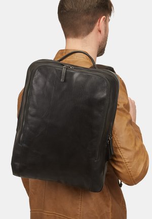 Sac à dos en cuir noir avec une texture lisse, présentant une forme rectangulaire et une poignée supérieure. Fermeture zippée et accents latéraux visibles.