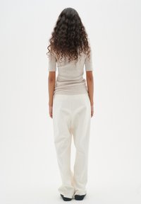 Hellbeige gestricktes Oberteil und lockere weiße Hose. Lockige Haare, von hinten zu sehen, mit langen Ärmeln und entspannter Passform aus weichem Stoff. Schwarze Schuhe.