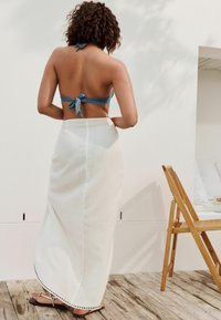 Blauwe halter bikini-top met strik aan de achterkant, witte linnen maxi-rok met een ronde zoom en zwarte stikselaccenten, gedragen met platte sandalen.
